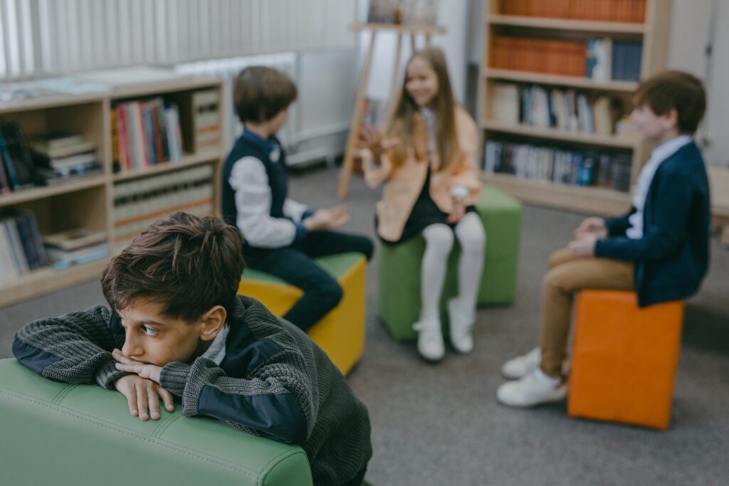 Sad boy is isolated from his classroom peers, while they chat with each other.