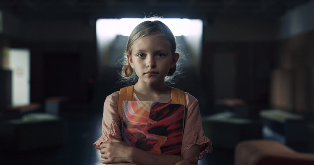 A girl standing in an empty and destroyed classroom holding a book.