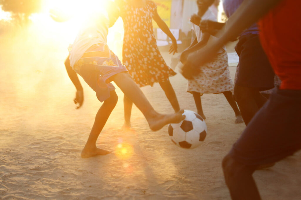 Children from various nationalities playing soccer in a dirt soccer field.