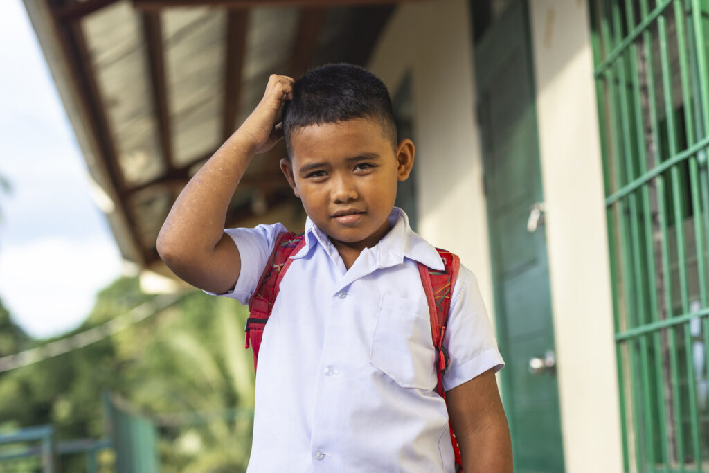 A young school boy scratches his head, not knowing the answer while being questioned. Outside a small rural school building.